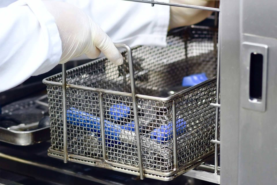 Person in a white lab coat and thick off-white gloves removing a metal mesh tray from the auto wash station at BoneSim.
