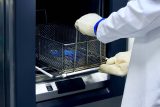 Person in a white lab coat and thick off-white gloves removing a metal mesh tray of instruments at BoneSim.